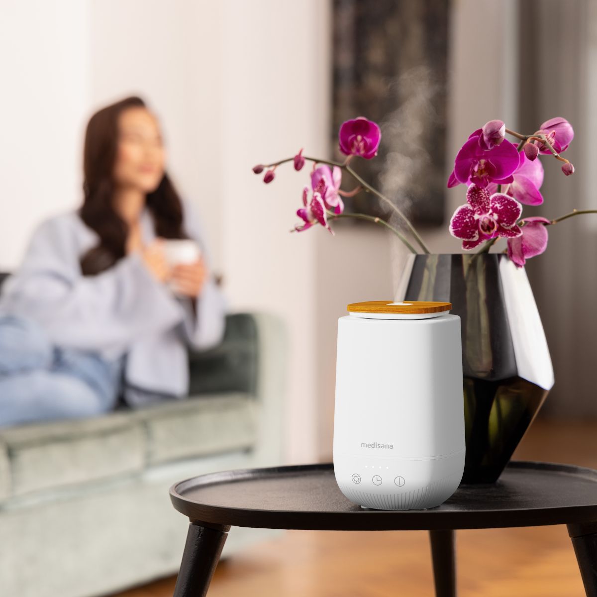 A white aroma diffuser stands on a table next to orchids while a woman relaxes in the background.